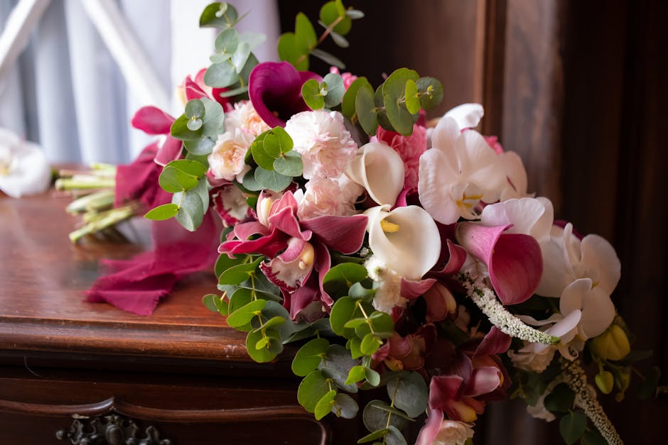 A vibrant floral bouquet featuring elegant white calla lilies, deep magenta anthuriums, soft pink roses, and sprigs of eucalyptus with rounded green leaves, arranged in a cascading style. The bouquet 