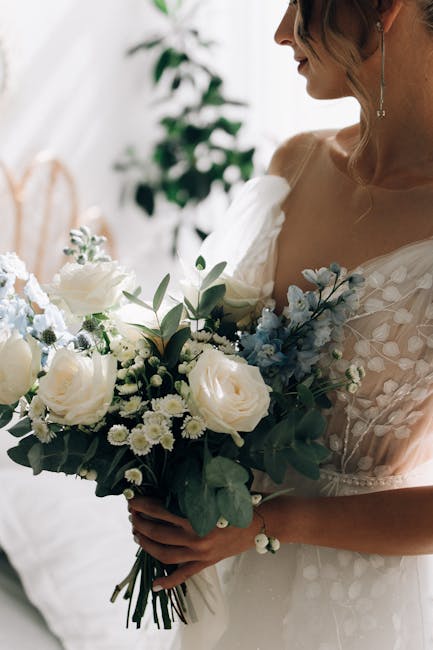 A bride holding a bouquet arrangement featuring cream and white roses, accented with small white filler flowers and green foliage, with hints of soft blue flowers. The bouquet is wrapped with a natura