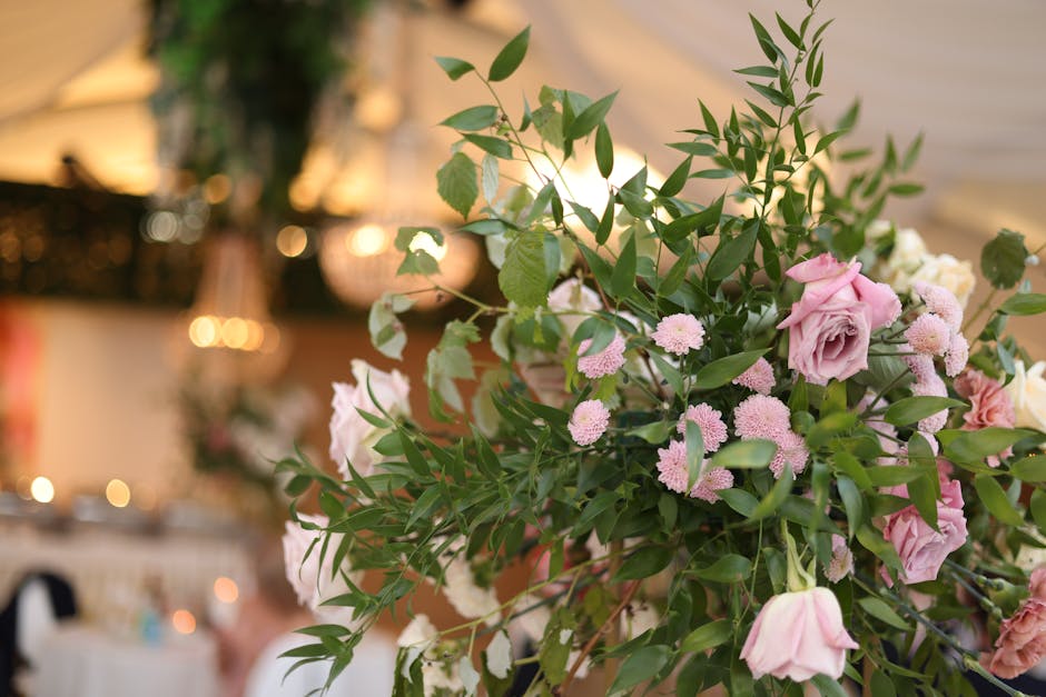 A large floral arrangement featuring pastel pink roses and small light pink chrysanthemum-like flowers, complemented by green foliage with elongated, glossy leaves. The bouquet is set against a softly