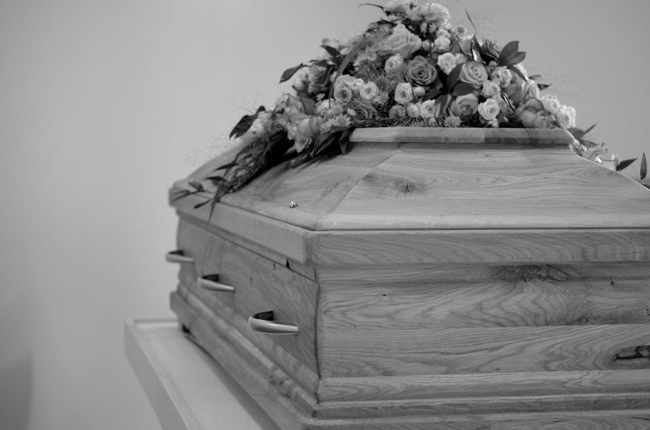 A wooden casket with a smooth, polished surface and natural wood grain, adorned with a floral arrangement consisting of roses, lilies, and other flowers in soft pastel colors, resting on a white table