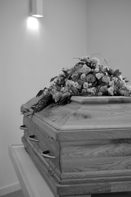 A detailed black and white photograph of a closed wooden coffin resting on a stand, topped with a large floral arrangement consisting of roses, lilies, and greenery. The bouquet appears freshly arrang