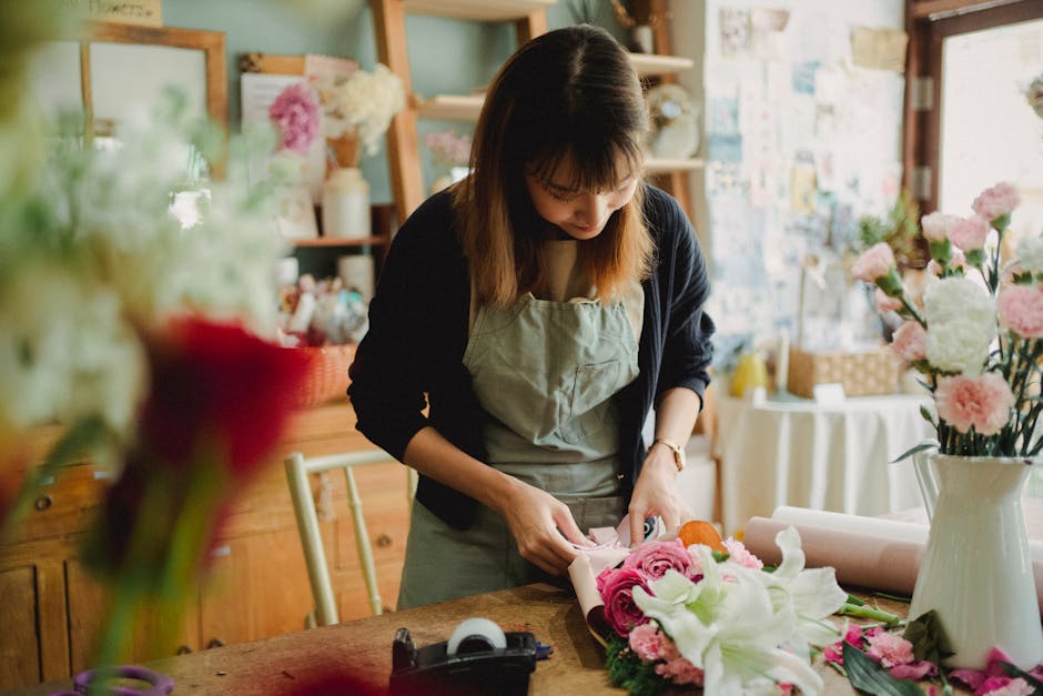 A florist in a cozy, well-lit flower shop, arranging a vibrant bouquet of pink and white roses, lilies, and greenery on a wooden table. She is wearing a beige apron over a black top and is focused on
