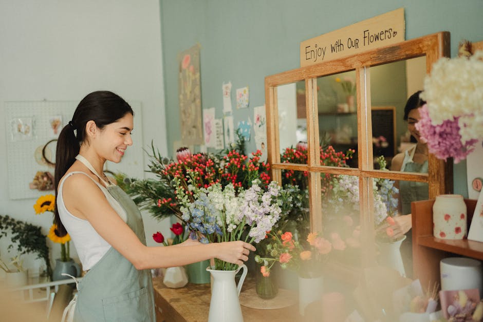 A young woman in a light-colored apron arranging a bouquet of fresh flowers inside a florist shop. She is smiling gently while holding a white ceramic pitcher filled with various flowers, including re