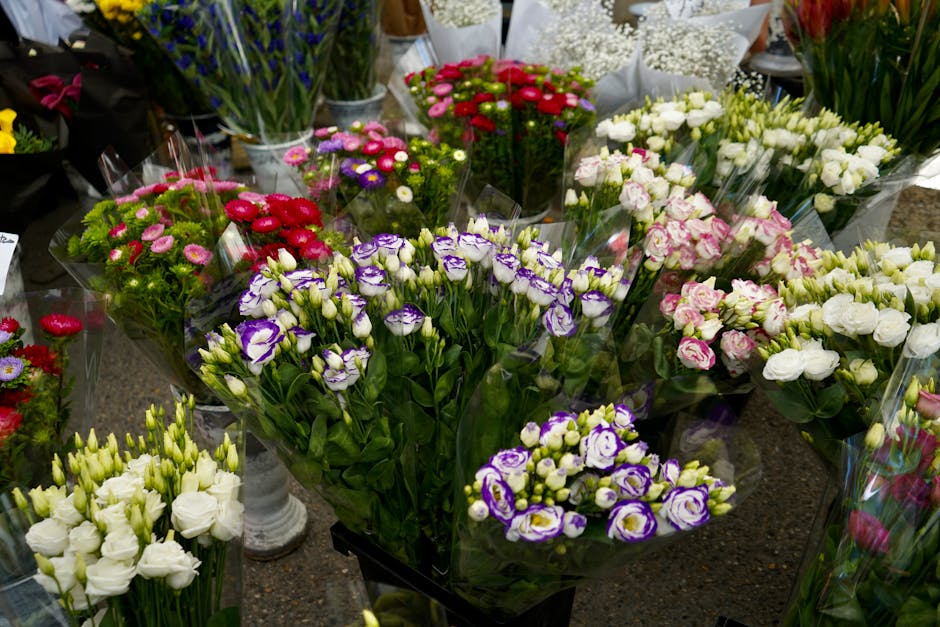 A display of fresh flower bouquets arranged on a dark surface, featuring various types of flowers wrapped in clear plastic. Prominent among them are clusters of purple and white lisianthus, white eust