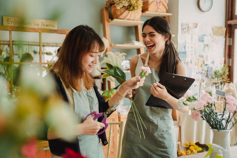 Two female florists inside a flower shop, one with shoulder-length brown hair and the other with long dark hair, are engaged in creating a floral gift. The woman with brown hair, dressed in a light gr