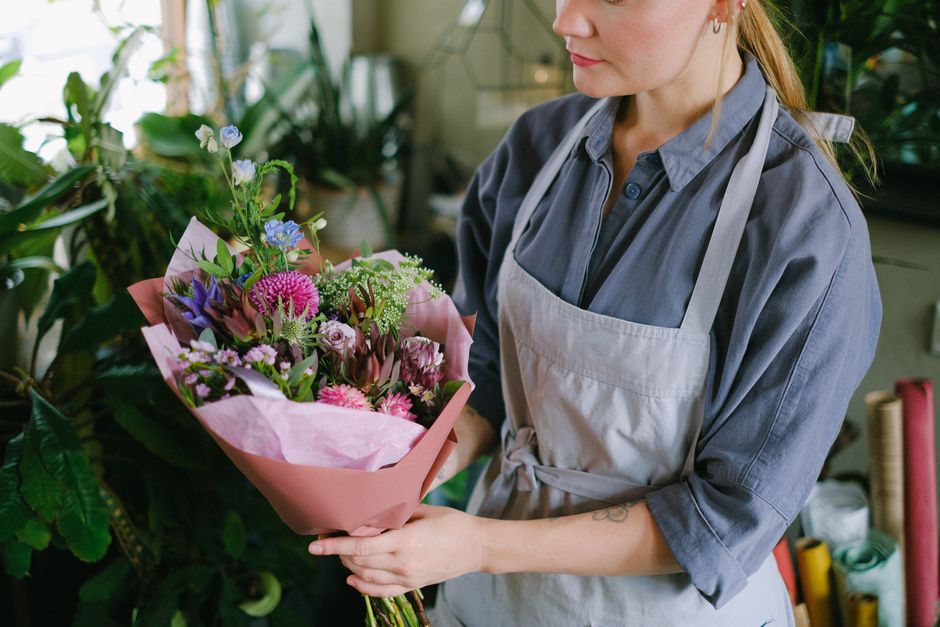 A woman in a grey button-up shirt and light grey apron holds a vibrant floral bouquet wrapped in pink tissue paper. The bouquet features a variety of fresh flowers, including pink and purple carnation